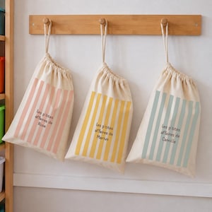 Può includere: Tre sacchetti color crema con coulisse appesi a un appendiabiti in legno. Ogni sacchetto presenta strisce verticali rosa, gialle o blu e il testo "Les p'tites affaires de" seguito dai nomi "Rose", "Marius" o "Camille" stampati in nero.