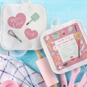 Peut inclure: Deux maniques blanches avec des motifs de cœurs roses et des autocollants d'ustensiles de cuisine. L'une porte les textes "Merci Celine" et "Noah". L'autre affiche une recette avec le texte "Recette de ma super nounou Celine".