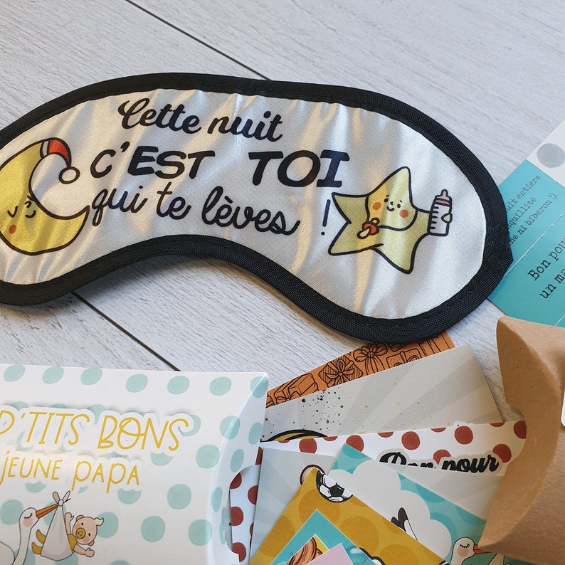 May include: A white sleep mask with black trim and a design of a yellow star holding a bottle and a crescent moon. The text on the mask reads "Cette nuit c'est toi qui te l&egrave;ves!"  A small box with blue and white polka dots reads "Petits Bons Jeune Papa".
