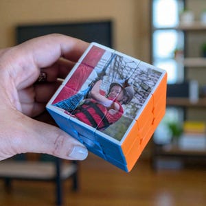 Op de afbeelding: Een gepersonaliseerde Rubik's Cube met een foto van twee personen. De kubus heeft blauwe en oranje zijden, met de fotosegmenten op de witte vierkanten. De afbeelding toont twee personen met helmen en rood-zwarte gestreepte shirts.