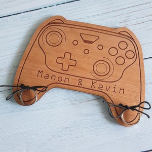 May include: A wooden game controller ring holder with the names "Manon & Kevin" engraved on it. Two rings are placed on the holder.