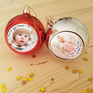 Peut inclure: Deux boules de Noël avec une photo d'un bébé dans un chapeau blanc et d'un bébé dans une robe rose. Les boules sont rouge et argent et portent le texte "Mon 1er Noël 2019".