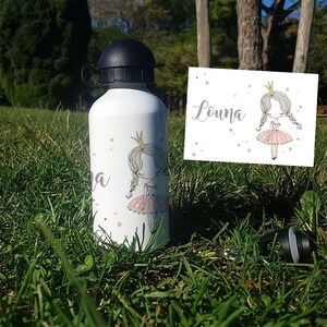 K&ouml;nnte beinhalten: Eine wei&szlig;e Wasserflasche mit einem rosa-wei&szlig;en Prinzessinnen-Design und dem Namen Louna. Die Flasche hat einen schwarzen Deckel und einen schwarzen Trageriemen.