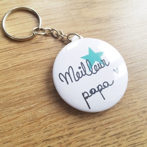 May include: White round keychain with a blue star and the text "Meilleur papa" in black cursive writing.