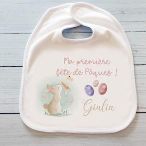 Pode incluir: Babador branco com um coelho rosa, três ovos de Páscoa pastel e o texto "Ma première fête de Pâques! Giulia".