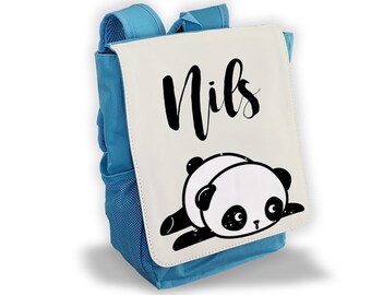Könnte beinhalten: Ein blauer Rucksack mit einer weißen Klappe mit dem Namen "Nils" in schwarzer Schrift und einer schwarz-weißen Panda-Illustration. Der Rucksack hat eine Seitentasche aus Mesh und blaue Träger.