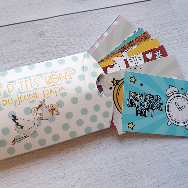 May include: A blue and white envelope with white polka dots and a stork carrying a baby. The envelope contains small cards with various illustrations and the text "Bon pour une grasse mat!"