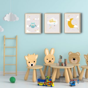 Könnte beinhalten: Drei gerahmte Drucke mit hellblauem Hintergrund und goldenen Akzenten. Die Drucke zeigen eine blaue Eule, den Namen "Arthur" in goldenen Buchstaben und einen gelben Halbmond. Die Drucke hängen an einer hellblauen Wand über einem Holztisch und vier Holzstühlen mit Tiergesichtern.