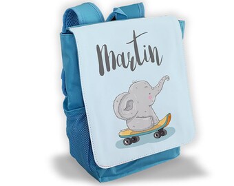 Könnte beinhalten: Ein blauer Rucksack mit einer hellblauen Frontklappe, auf der ein Cartoon-Elefant auf einem Skateboard zu sehen ist. Die Klappe trägt auch den Namen "Martin" in schwarzer Kursivschrift.