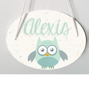 Könnte beinhalten: Ein weißes ovales Holzschild mit einer hellgrünen Eule und dem Namen "Alexis" in hellgrüner kursivschrift. Das Schild hat einen weißen Hintergrund mit hellgelben und hellblauen Farbspritzern.