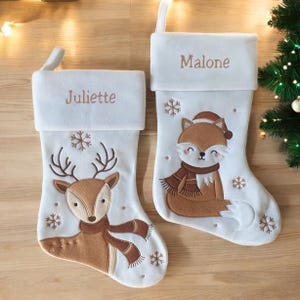 Könnte beinhalten: Zwei weiße Weihnachtsstrümpfe mit braunen Tierstickereien. Ein Strumpf zeigt ein Reh mit Schal und dem Namen Juliette, der andere einen Fuchs mit Schal und Mütze sowie dem Namen Malone, beide verziert mit kleinen Schneeflocken-Stickereien.