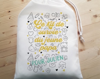 以下が含まれることがあります： 黄色と黒のベビー関連のアイコンと「Le Kit de survie du jeune papa POUR JULIEN」というテキストが描かれた白い巾着袋です。