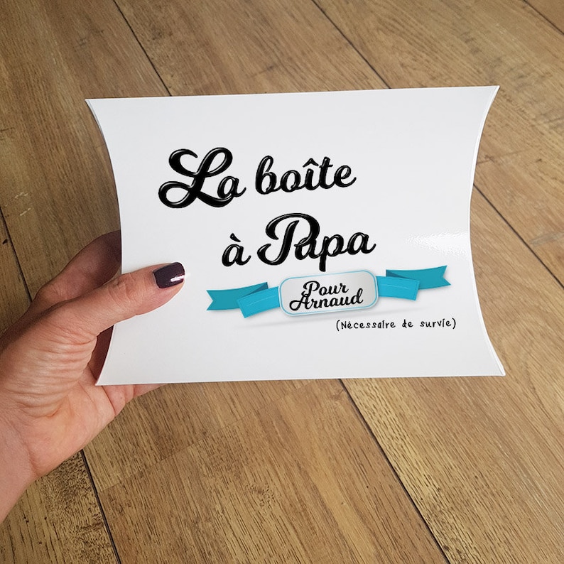 May include: White gift box with the text "La bo&icirc;te &agrave; Papa" in black cursive lettering. A blue ribbon with the text "Pour Arnaud" is below the text. The text "(N&eacute;cessaire de survie)" is below the ribbon.