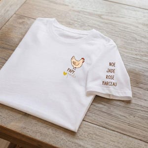 Può includere: Una t-shirt bianca a maniche corte con una piccola illustrazione di una gallina marrone chiaro sopra il testo "PAPY poule" e un cuore giallo. Sulla manica sono stampati in marrone i nomi "NOE", "JADE", "ROSE" e "MARCEAU".