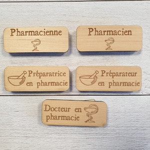 Peut inclure: Cinq étiquettes de nom en bois avec différents titres liés à la pharmacie. Les étiquettes présentent un mortier et un pilon, un serpent enroulé autour d'une coupe, et les mots "Pharmacienne", "Pharmacien", "Préparatrice en pharmacie", "Préparateur en pharmacie", et "Docteur en Pharmacie".