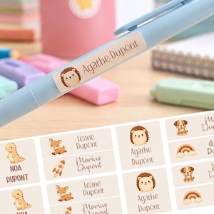 May include: A light blue plastic pen featuring a rectangular adhesive label with a cartoon hedgehog illustration and the printed name Agathe Dupont. Below, a sheet of matching rectangular name labels displays various cartoon icons.