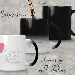 Può includere: Una tazza magica nera con un cuore rosa e il testo "Mon coeur je taime depuis 1254 jours" che appare quando si versa un liquido caldo nella tazza. La tazza è su una superficie grigia. Il testo "Surprise..." è scritto sopra la tazza. Il testo "...le message apparait avec la chaleur" è scritto sotto la tazza.