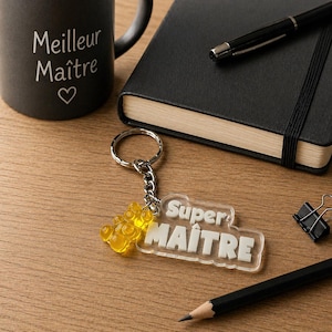 Op de afbeelding: Een zwarte mok met de tekst "Meilleur Maître" en een hartsymbool. Een doorzichtige sleutelhanger met de woorden "Super MAÎTRE" en een gele pootvormige bedel. Een zwart notitieboek, pen en potlood liggen ook op het houten oppervlak.