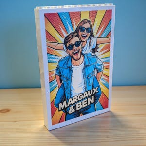 Può includere: Un blocco bianco con un'illustrazione in stile fumetto di una coppia con occhiali da sole, con il testo "MARGAUX & BEN". Lo sfondo presenta un'esplosione di raggi rossi, gialli e blu.