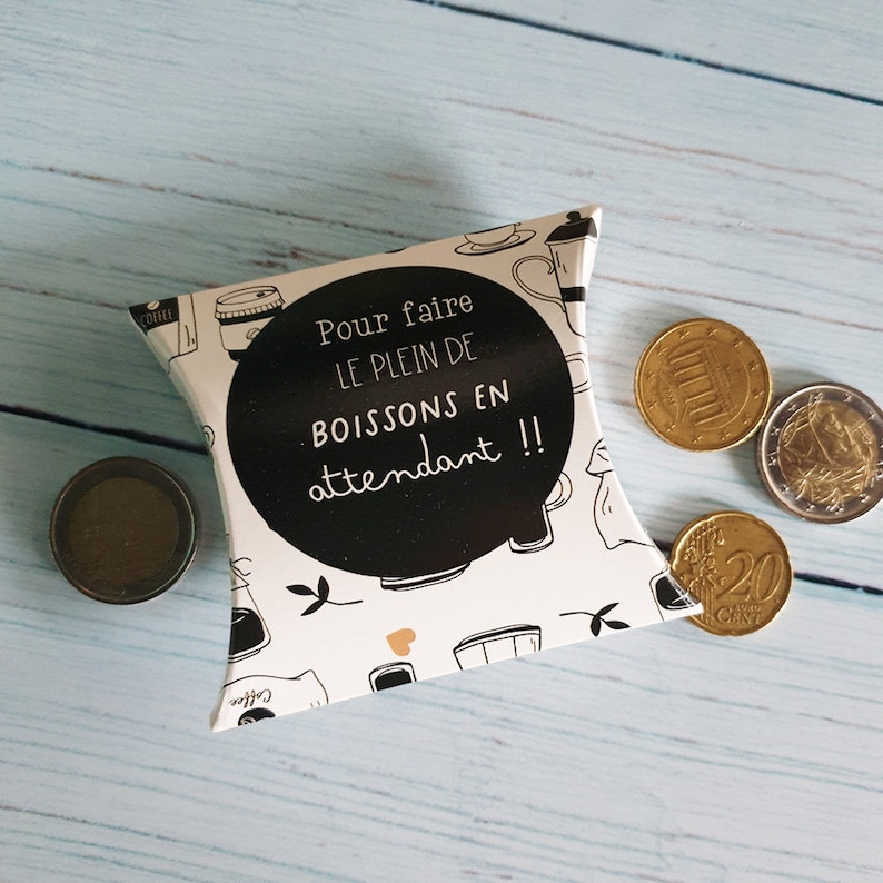 May include: A white and black pillow-shaped box with a black and white design of coffee cups and the text "Pour faire le plein de boissons en attendant!!". The box is sitting on a light blue wooden surface with four gold coins.
