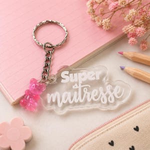 Op de afbeelding: Een heldere acryl sleutelhanger met de woorden "Super maitresse" in witte letters. De sleutelhanger heeft een zilveren ketting en ring, en een roze gummybeer bedel.
