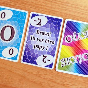 Pode incluir: Três cartas de jogo retangulares com cantos arredondados. A carta da esquerda é azul com um padrão de favo de mel e o número zero. A carta central é roxa com um padrão de favo de mel, o número menos dois e o texto Bravo! Tu vas être papy!. A carta da direita apresenta um padrão de favo de mel com gradiente de arco-íris e a palavra SKYJO.
