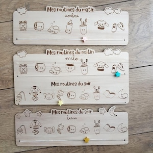 May include: Three wooden boards with a routine chart for children. Each board has a different name at the top: Ambre, Milo, and Liam. The boards have images of a clock, a toothbrush, a cup, a book, a shower, and other items.