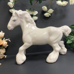 May include: A white ceramic horse figurine with a flowing mane and tail. The horse is in a trotting pose. The figurine is glossy and has a smooth surface. Decorative flowers and greenery are in the background.