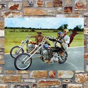 May include: Three people riding on two motorcycles on a paved road. The motorcycles are chrome and have a vintage look. The people are wearing helmets and leather jackets. The road is surrounded by green grass and trees.