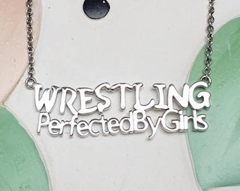 Op de afbeelding: Een zilverkleurige ketting met het woord "WRESTLING" boven "PerfectedByGirls" in een gestileerd lettertype. De ketting hangt aan een fijne ketting. De achtergrond is een kleurrijk, abstract ontwerp met zwarte stippen.