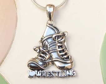 Op de afbeelding: Zilverkleurige hangerketting met een paar worstelschoenen. De hanger heeft het woord "WRESTLING" onder de schoenen. De ketting wordt getoond op een witte en pastelkleurige achtergrond.