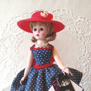 May include: A vintage doll wearing a red hat with a floral accent and a navy blue polka dot dress. The doll has a pearl necklace and red shoes. A tag reads "Vintage Madame Alexander".