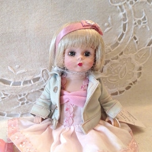 May include: A porcelain doll wearing a pink dress with a denim jacket, a pink headband, and pink shoes. The doll has blonde hair and blue eyes.
