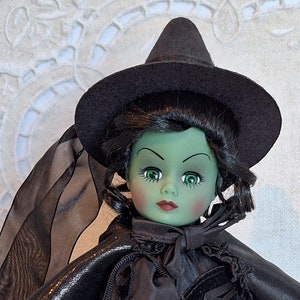 Puede incluir: Muñeca de bruja con piel verde, pelo negro y sombrero negro. La muñeca viste un vestido negro y tiene un velo negro. La muñeca tiene ojos verdes y labios rojos. Un artículo de colección.