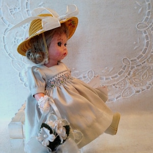 May include: A porcelain doll wearing a white dress and a yellow straw hat with a white ribbon. The doll is holding a bouquet of white flowers and is sitting on a white lace tablecloth.