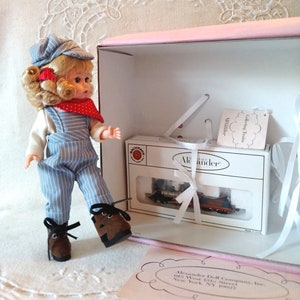 May include: A doll wearing blue and white striped overalls, a red bandana, and a blue cap. The doll is holding a small toy train set in a white box with the words "Alexander" and "Collecting Trains" printed on it.