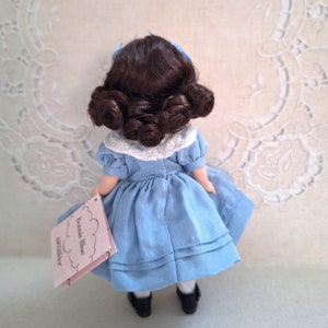 Puede incluir: Una peque&ntilde;a mu&ntilde;eca de porcelana que lleva un vestido azul con un cuello de encaje blanco. La mu&ntilde;eca tiene el pelo casta&ntilde;o recogido en dos mo&ntilde;os y sostiene una etiqueta que dice "Jennie Blue" y "Alexander".