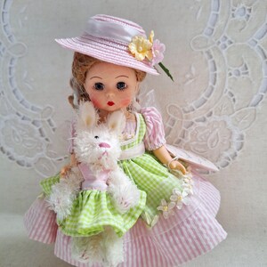 May include: A vintage doll wearing a pink and green gingham dress, a matching hat, and holding a white stuffed bunny. The doll's dress has a pink and white striped skirt. The hat is decorated with flowers.