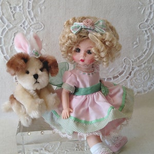 May include: A vintage doll with blonde curly hair, wearing a pink dress with green trim, and a matching bow. A stuffed puppy with bunny ears sits beside the doll. The doll and puppy are on a clear acrylic box.