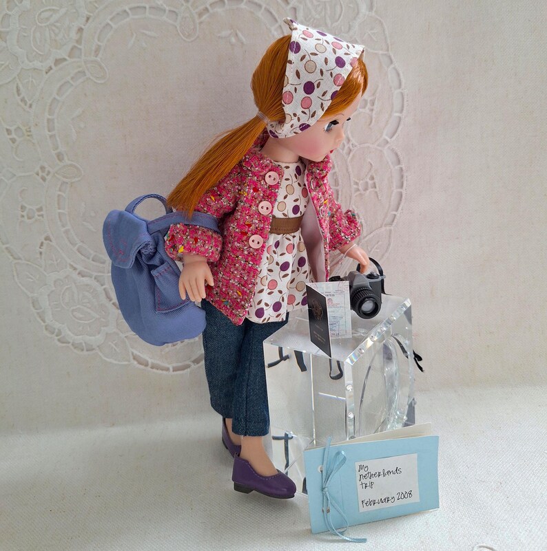 May include: A doll wearing a pink and white patterned jacket, a blue backpack, and a blue and white patterned headband. The doll is holding a camera and a passport. The doll is standing on a clear acrylic block. The text on the card reads "My Memories &  Travels February 2018"