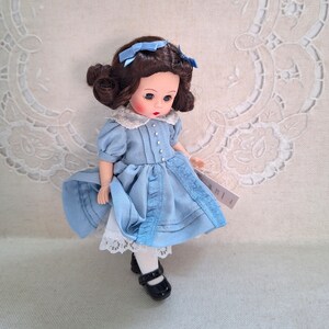 Puede incluir: Una mu&ntilde;eca de porcelana con cabello casta&ntilde;o y ojos azules, vestida con un vestido azul claro con ribete de encaje blanco y zapatos negros.