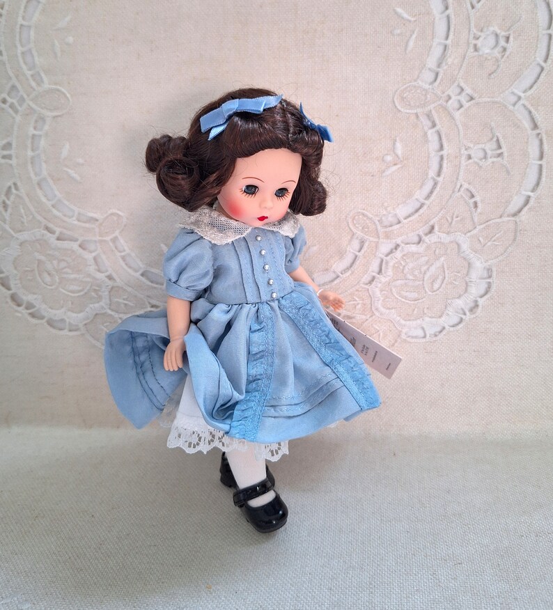 Puede incluir: Una mu&ntilde;eca de porcelana con cabello casta&ntilde;o y ojos azules, vestida con un vestido azul claro con ribete de encaje blanco y zapatos negros.