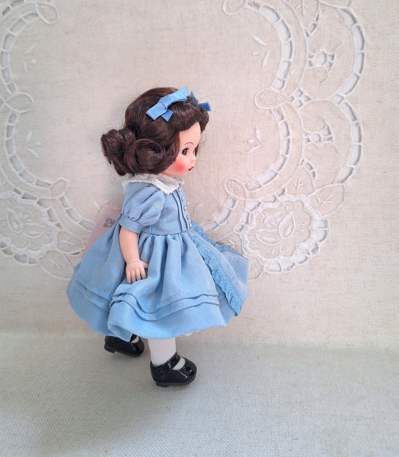 Puede incluir: Una mu&ntilde;eca de porcelana con cabello casta&ntilde;o que lleva un vestido azul claro y zapatos negros. La mu&ntilde;eca est&aacute; de pie sobre una superficie blanca con un patr&oacute;n de encaje.