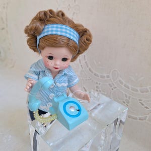 May include: A vintage doll with auburn hair and a blue gingham headband and shirt, holding a light blue rotary phone. The doll has rosy cheeks and is standing next to a clear, glass-like block.