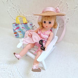 May include: A vintage doll in a pink gingham swimsuit and straw hat, seated in a white beach chair. The doll holds black sunglasses and is next to a blue tote bag with a fish pattern. The scene evokes a summer beach theme.