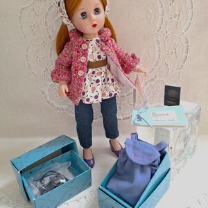 May include: A red-haired doll wearing a pink and white patterned jacket, a white and purple patterned blouse, blue jeans, and a purple headband. The doll is holding a blue bag and is standing on a white surface. The doll is part of a set that includes a blue bag, a pair of shoes, and a small box.