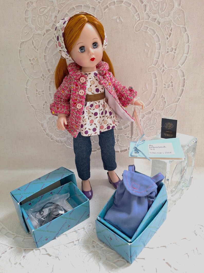 May include: A red-haired doll wearing a pink and white patterned jacket, a white and purple patterned blouse, blue jeans, and a purple headband. The doll is holding a blue bag and is standing on a white surface. The doll is part of a set that includes a blue bag, a pair of shoes, and a small box.