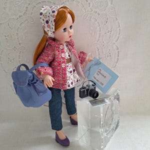 May include: A red-haired doll wearing a pink and white patterned jacket, a white and pink patterned hat, blue jeans, and purple shoes. The doll is holding a blue and white note that says "Paris Hotel Visit" and a small camera. The doll is also carrying a blue backpack.