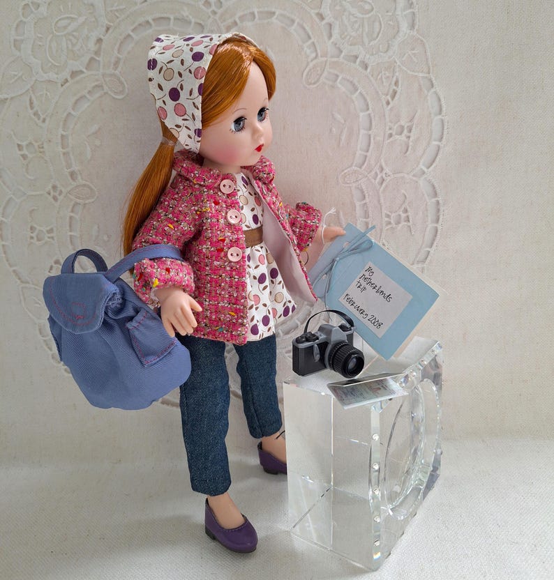 May include: A red-haired doll wearing a pink and white patterned jacket, a white and pink patterned hat, blue jeans, and purple shoes. The doll is holding a blue and white note that says "Paris Hotel Visit" and a small camera. The doll is also carrying a blue backpack.