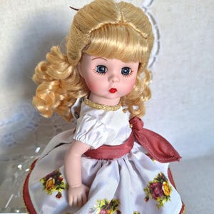 Könnte beinhalten: Eine Vintage-Porzellanpuppe mit blonden Locken und blauen Augen. Die Puppe trägt ein weißes Kleid mit bordeauxfarbenem Schärpe und Blumendruck. Das Kleid hat goldfarbene Verzierungen und eine passende goldfarbene Halskette.
